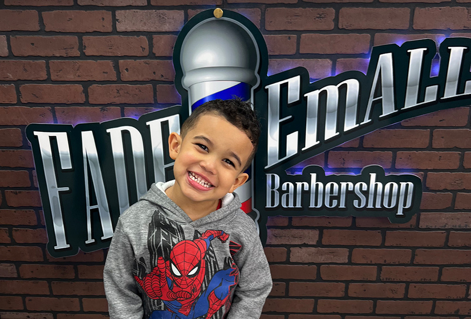 Smiling young client in front of the lit Fade 'Em All Barbershop sign