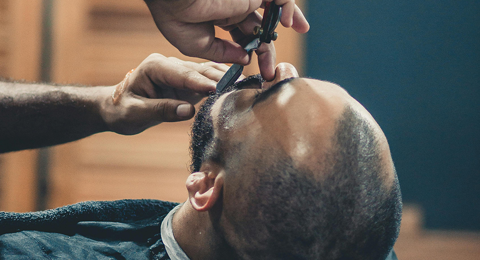 Barber lining up a reclined client's mustache with a straight razor